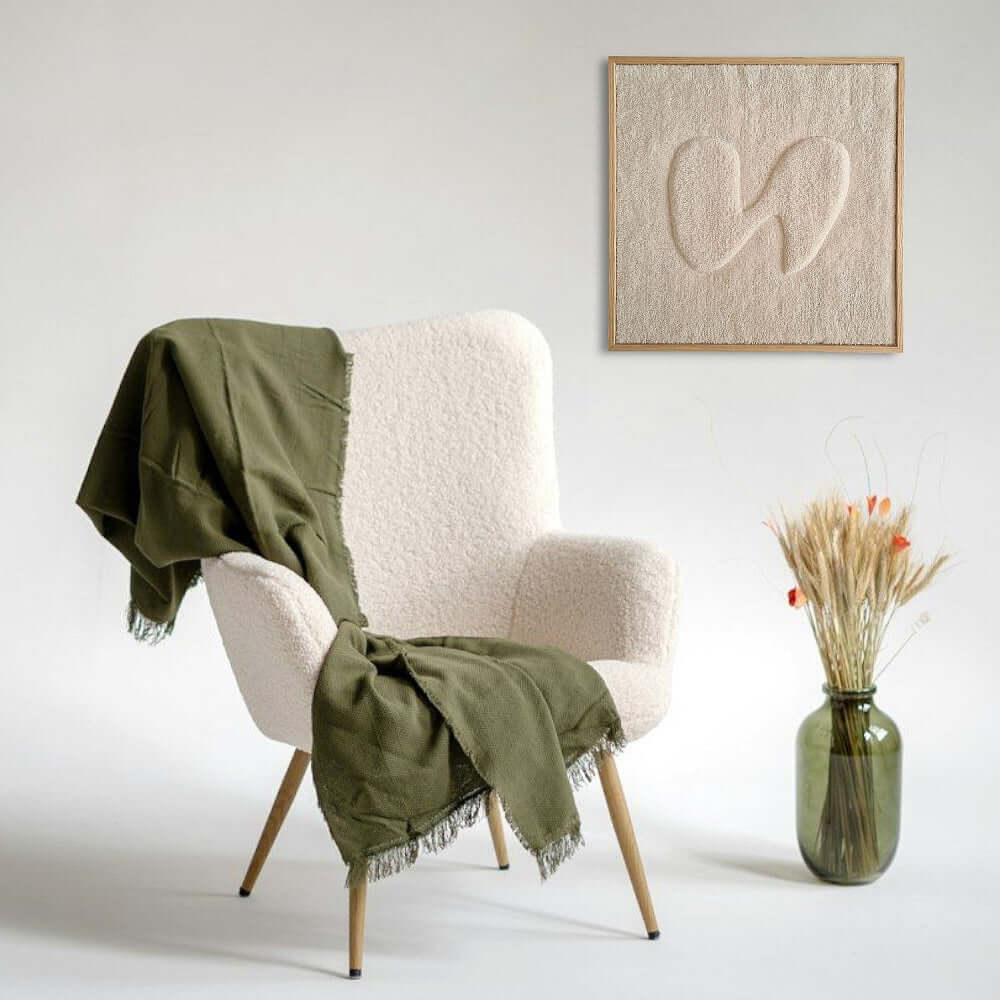 handgewebter, gerahmter Wandteppich in einen Echtholzrahmen in Eiche natur in der Größe 55x55 cm, gerahmter Wandteppich in beige mit organischer Form in 3D-Optik in hellem Raum mit Teddyfell bezogenem Sessel und olivgrüner Kuscheldecke